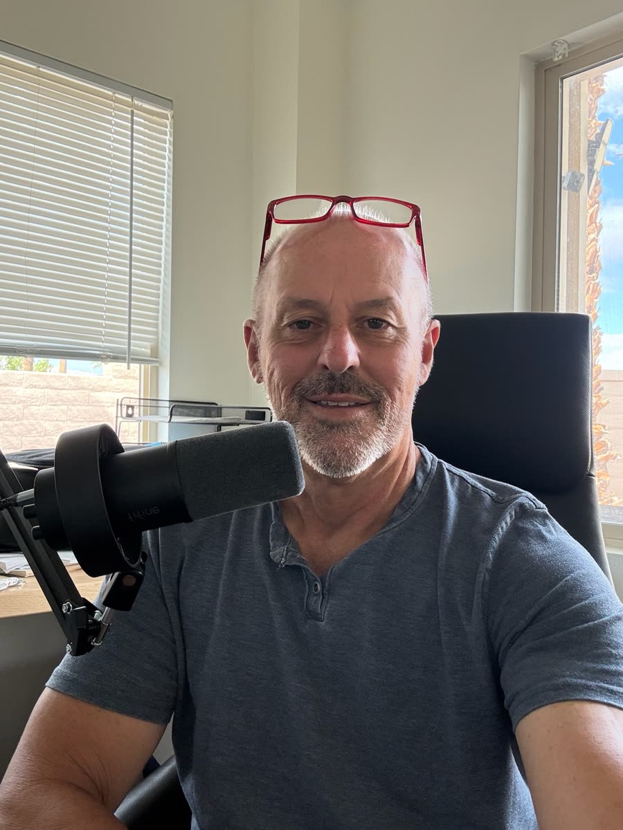 Alan smiling at his desk with a podcast microphone, red reading glasses on his head